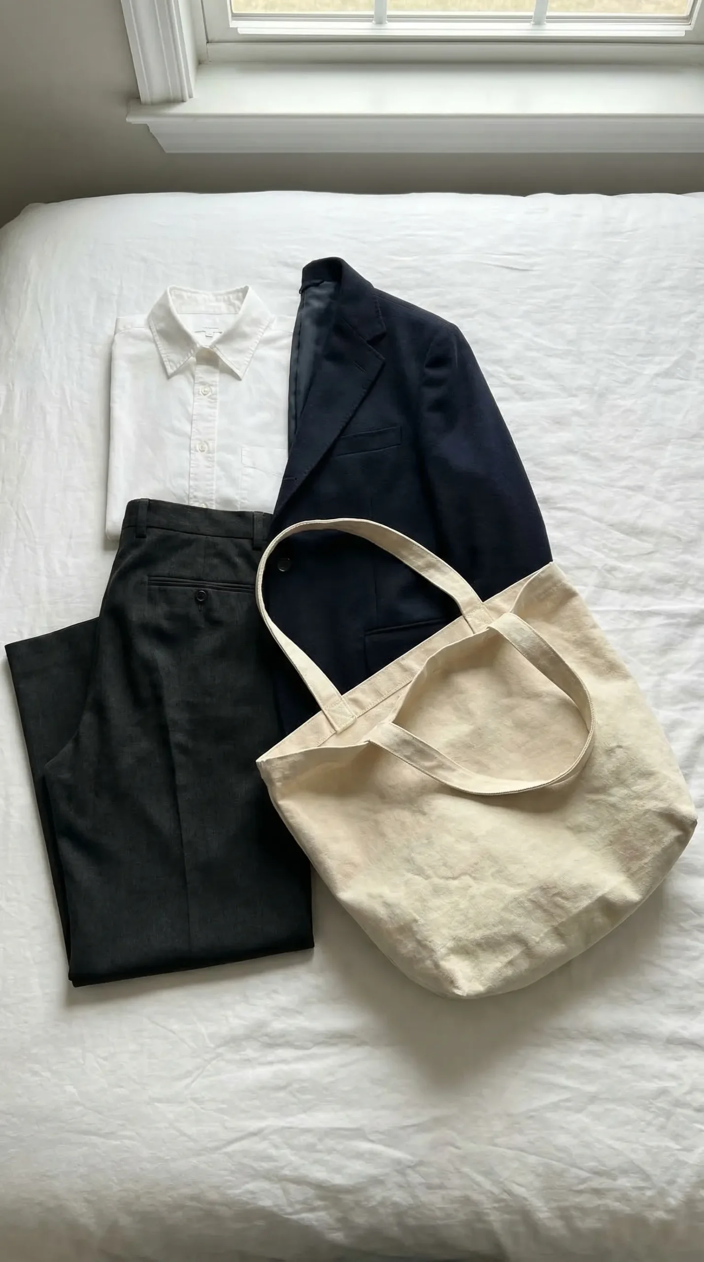 Flat lay on a bed showing a white shirt, navy blazer and dark trousers next to a canvas tote — the formality mismatch between sharp clothes and a casual bag is immediately visible