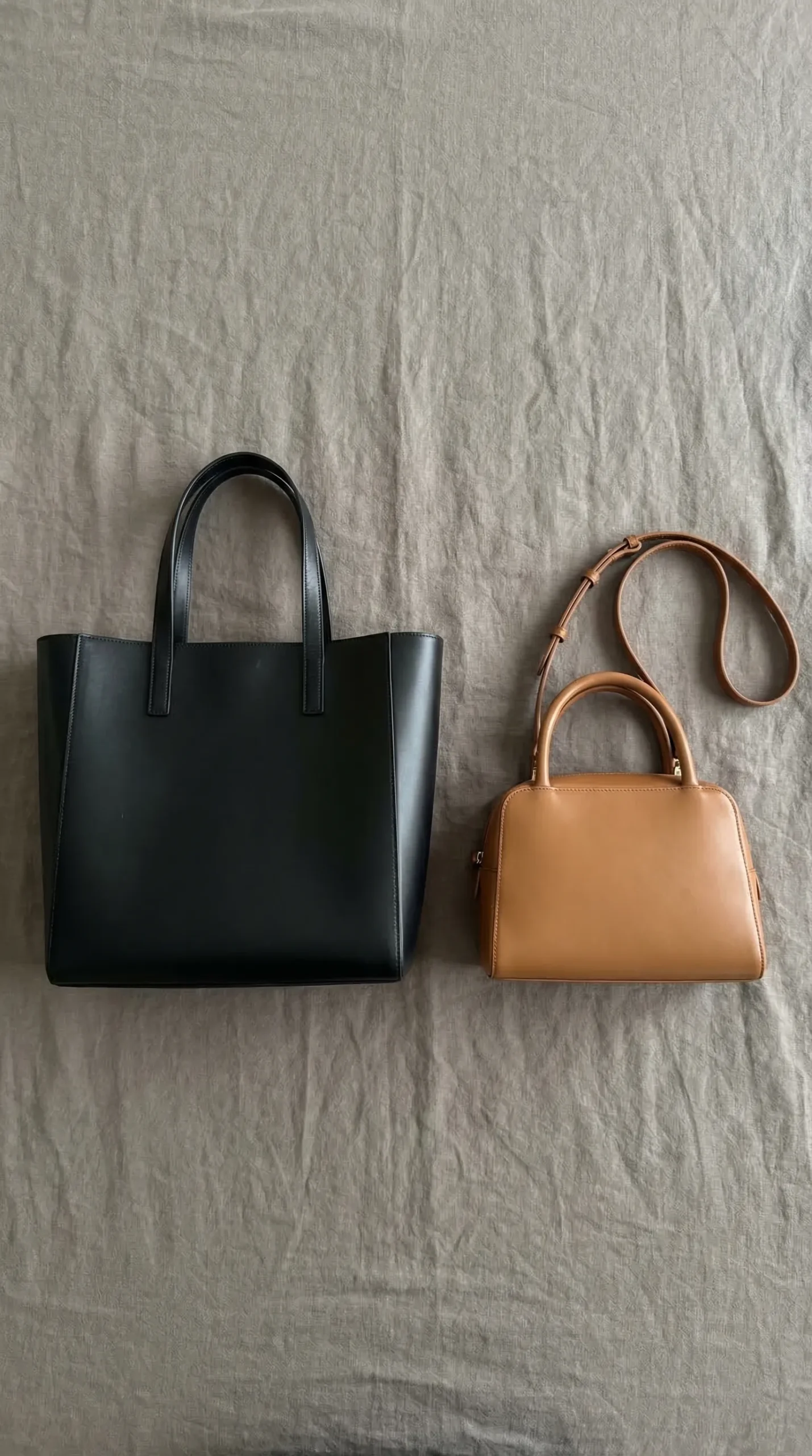Flat lay of two bags side by side — a large structured black leather tote on the left for transport, a smaller tan top-handle shoulder bag on the right for meetings