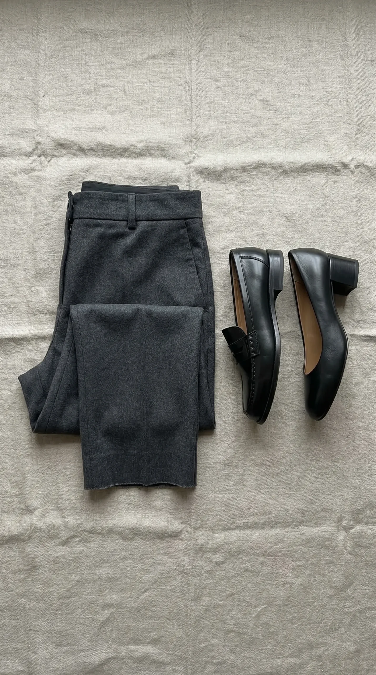 Flat lay of cropped straight-leg grey trousers next to a black loafer and black block heel — one trouser length works with both shoes