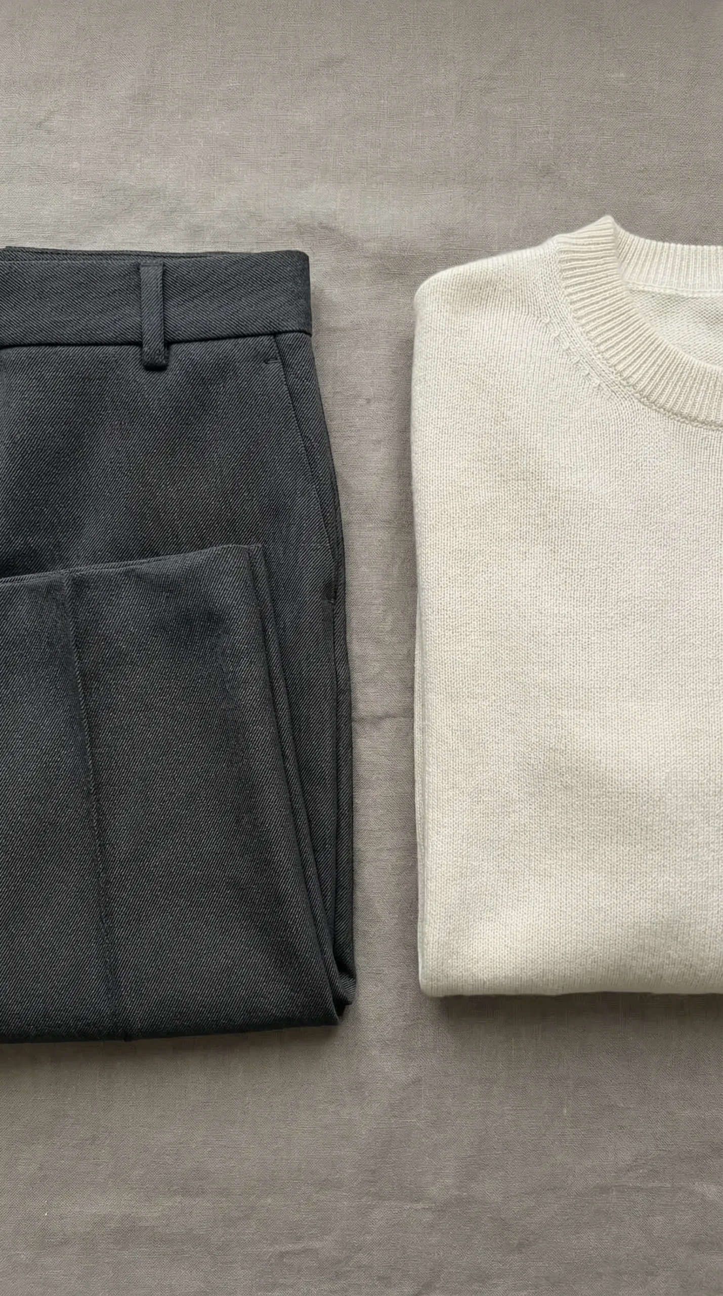 Flat lay of two anchor pieces — charcoal wide-leg twill trousers and a cream flat-knit merino crewneck