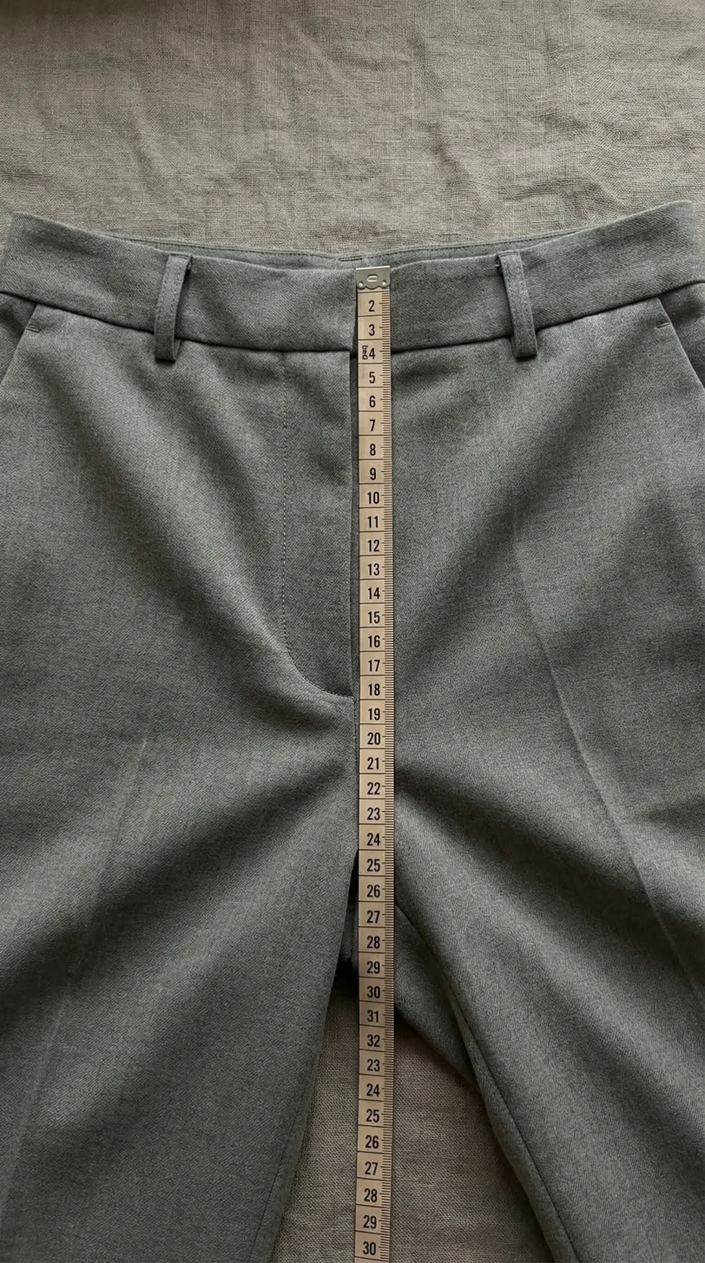 Flat lay of grey tailored trousers with measuring tape showing the front rise measurement from crotch seam to waistband